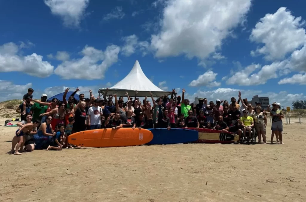 Imagem de participantes do projeto Abba Surf desfrutando de atividades aquáticas inclusivas na orla do Balneário Rincão, com instrutores e voluntários auxiliando pessoas com deficiência em pranchas de stand-up paddle, promovendo lazer e inclusão social