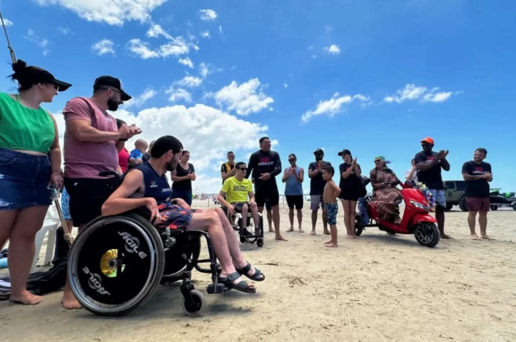 Imagem de participantes do projeto Abba Surf desfrutando de atividades aquáticas inclusivas na orla do Balneário Rincão, com instrutores e voluntários auxiliando pessoas com deficiência em pranchas de stand-up paddle, promovendo lazer e inclusão social