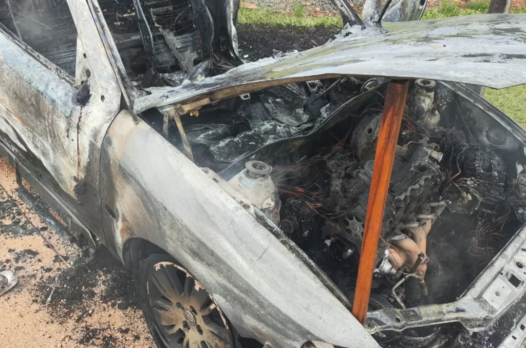 Carro completamente destruído após incêndio, com a lataria queimada e bombeiros no local realizando o rescaldo das chamas