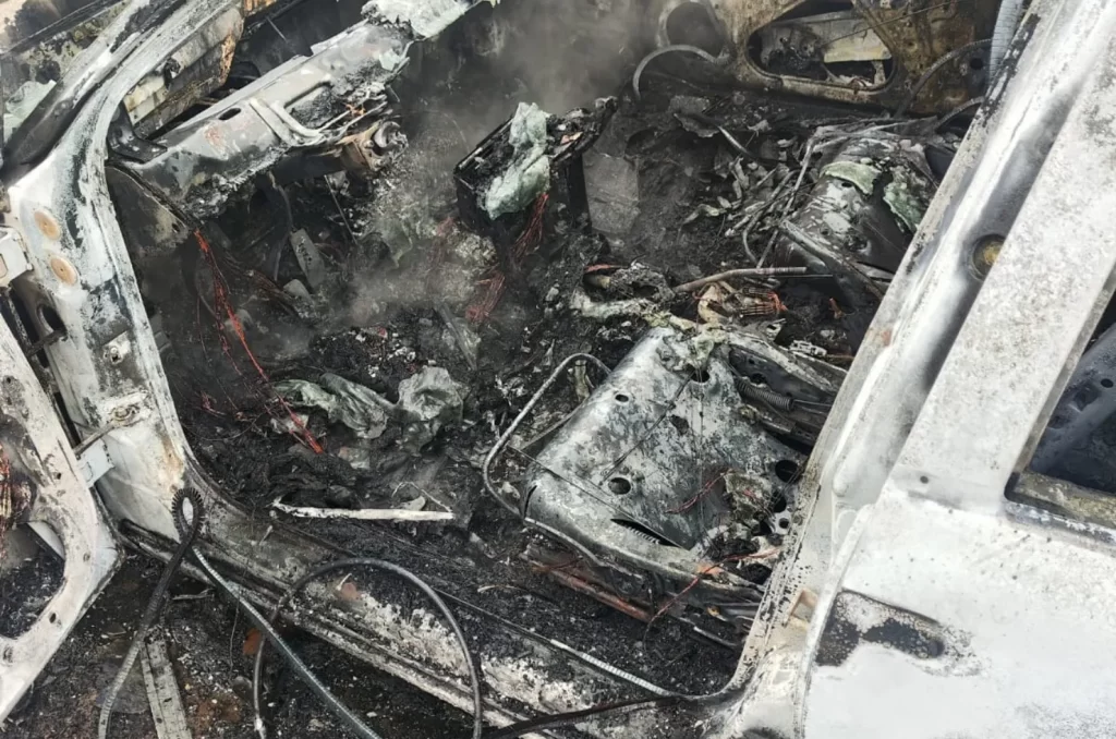 Carro completamente destruído após incêndio, com a lataria queimada e bombeiros no local realizando o rescaldo das chamas