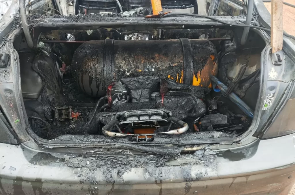 Carro completamente destruído após incêndio, com a lataria queimada e bombeiros no local realizando o rescaldo das chamas