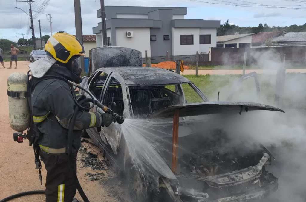 Carro completamente destruído após incêndio, com a lataria queimada e bombeiros no local realizando o rescaldo das chamas