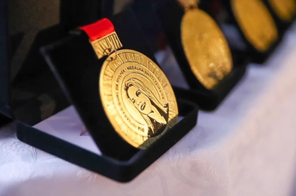 30 mulheres recebendo a Medalha Thati Teixeira em solenidade no jardim interno do Paço Municipal Marcos Rovaris, em Criciúma