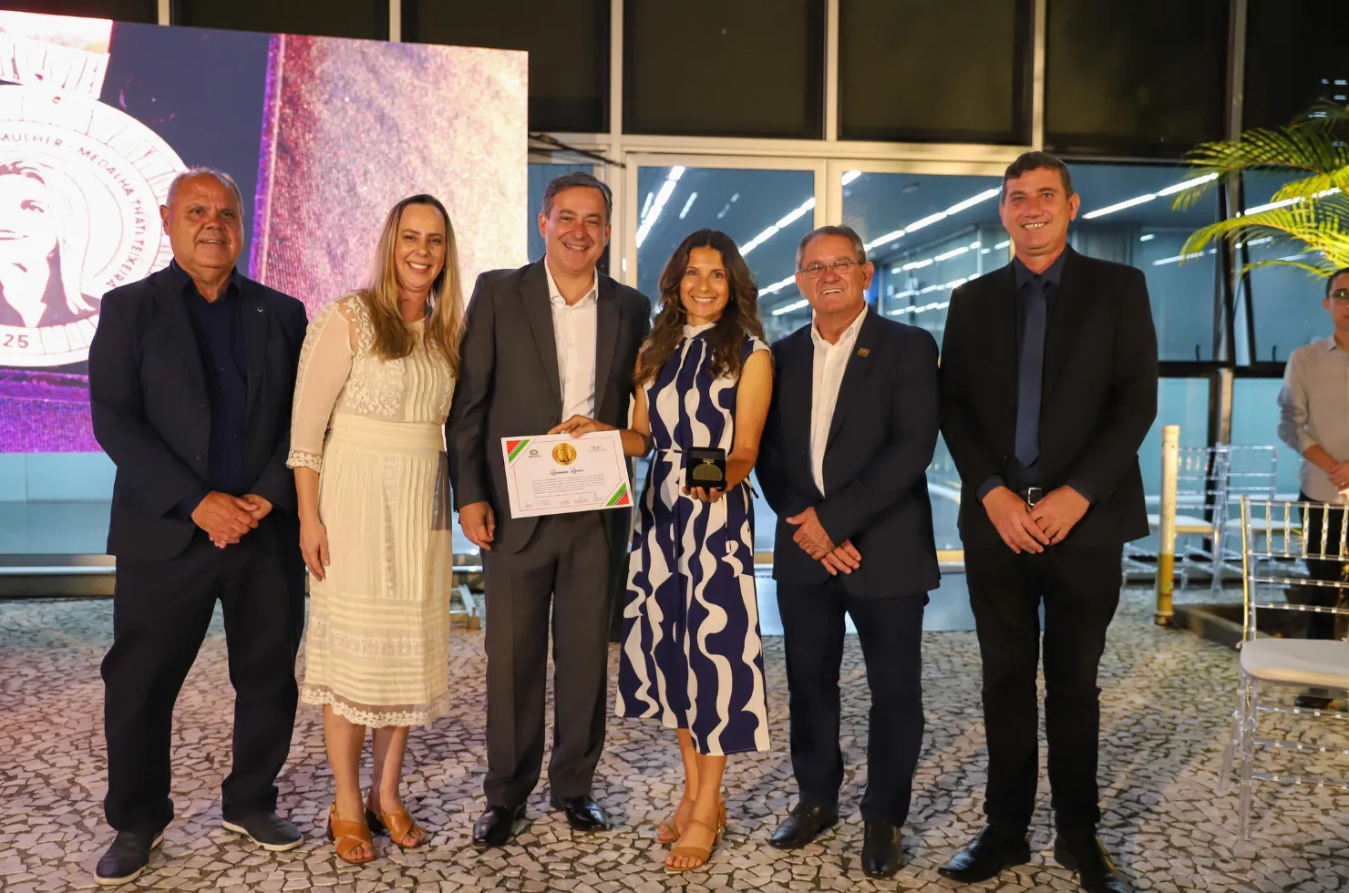 30 mulheres recebendo a Medalha Thati Teixeira em solenidade no jardim interno do Paço Municipal Marcos Rovaris, em Criciúma