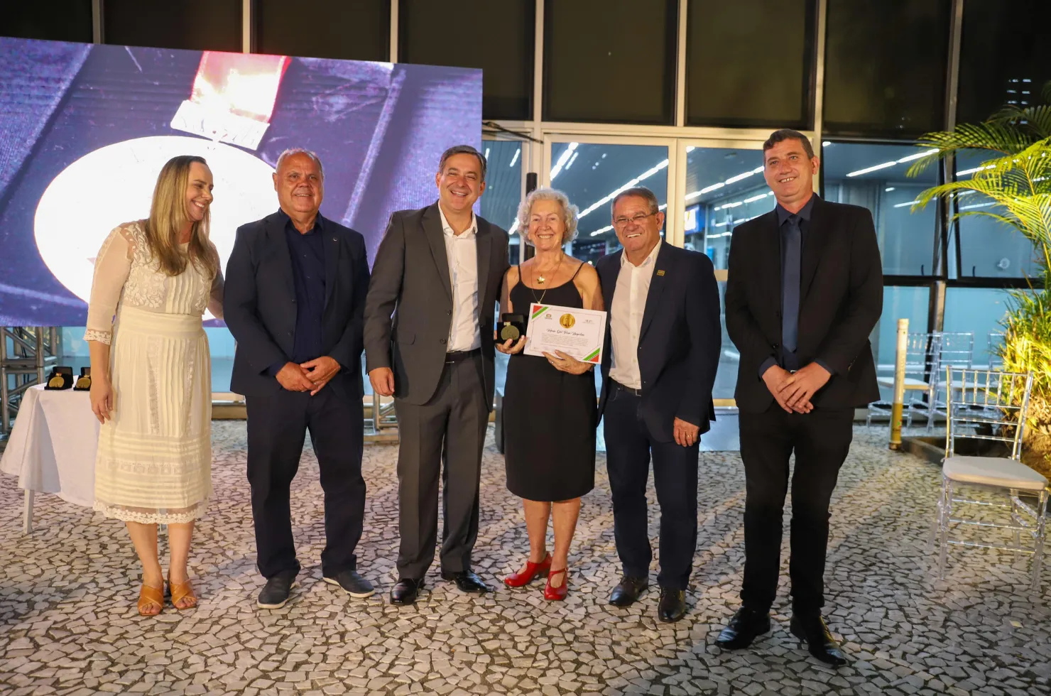 30 mulheres recebendo a Medalha Thati Teixeira em solenidade no jardim interno do Paço Municipal Marcos Rovaris, em Criciúma