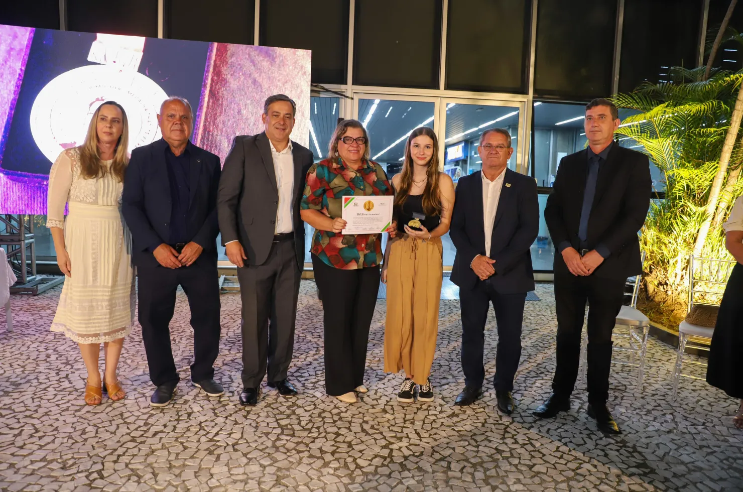 30 mulheres recebendo a Medalha Thati Teixeira em solenidade no jardim interno do Paço Municipal Marcos Rovaris, em Criciúma