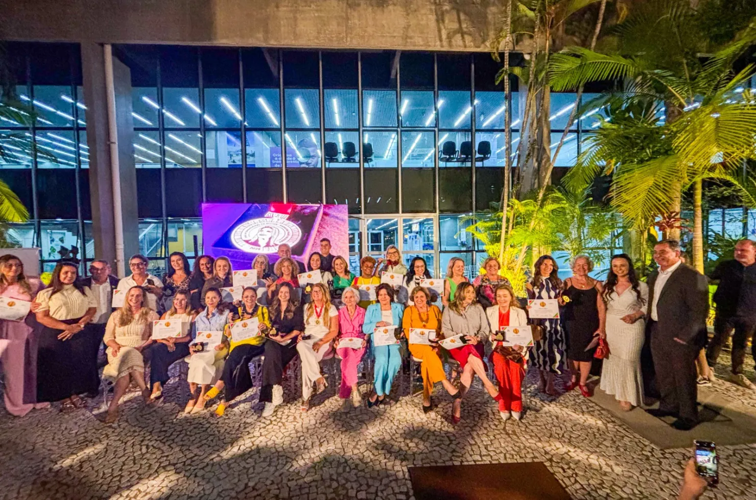 30 mulheres recebendo a Medalha Thati Teixeira em solenidade no jardim interno do Paço Municipal Marcos Rovaris, em Criciúma