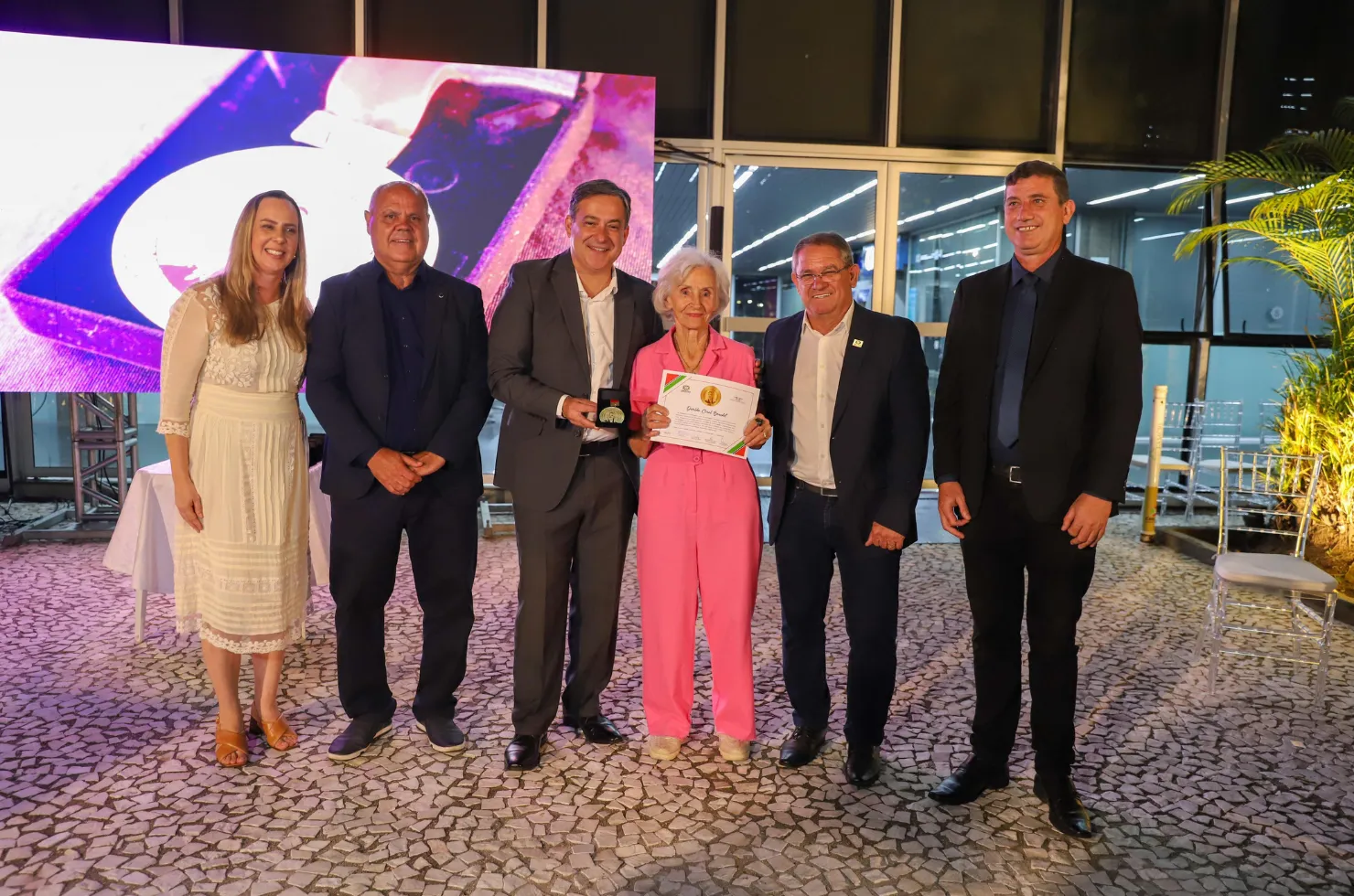 30 mulheres recebendo a Medalha Thati Teixeira em solenidade no jardim interno do Paço Municipal Marcos Rovaris, em Criciúma