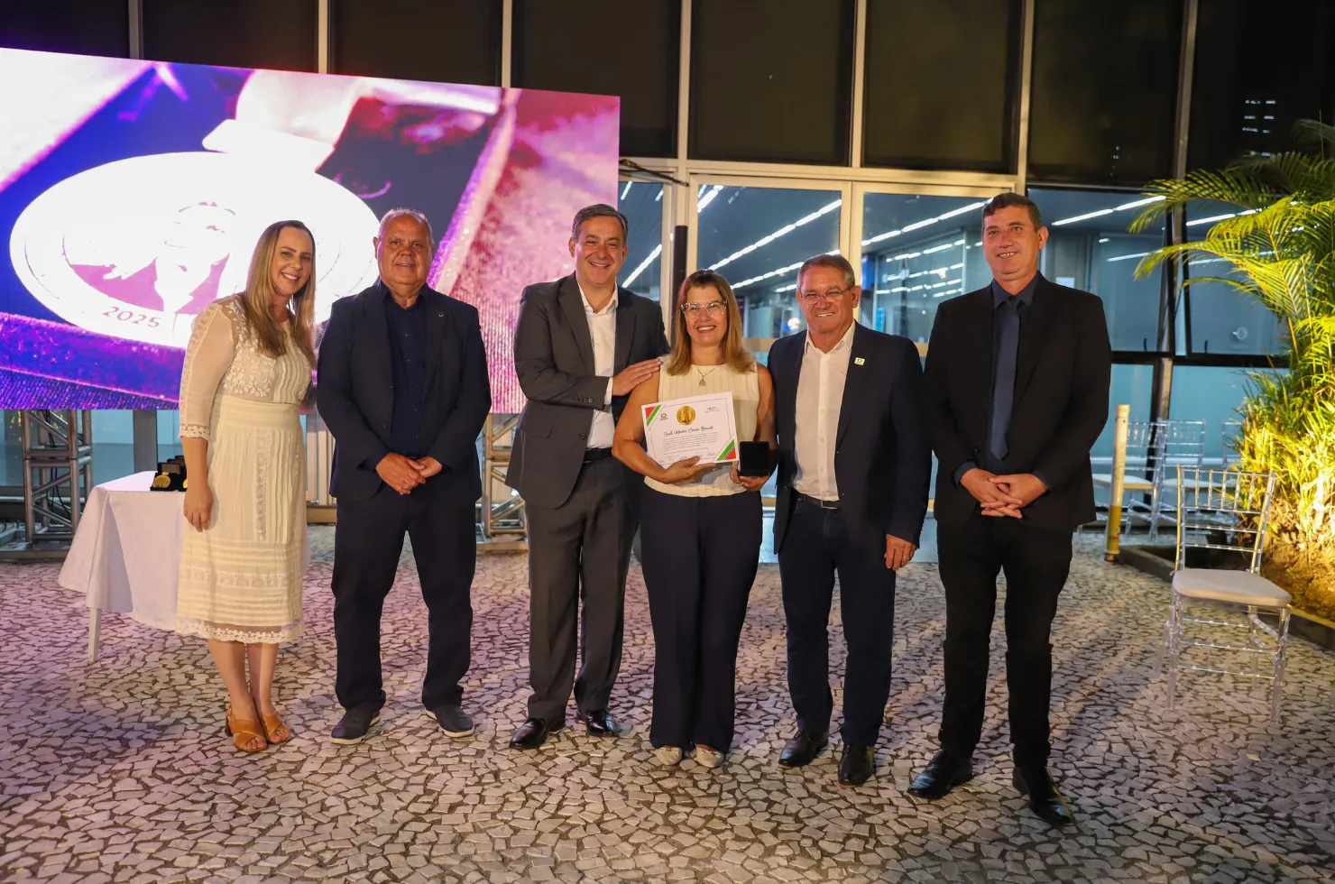 30 mulheres recebendo a Medalha Thati Teixeira em solenidade no jardim interno do Paço Municipal Marcos Rovaris, em Criciúma